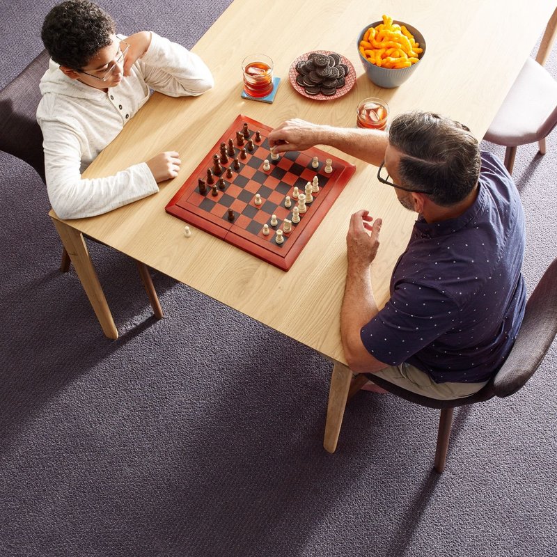father and son playing chess and eating snacks on a brown table in a living room with gray carpet from The Carpet and Drapery Shoppe in Escanaba, MI